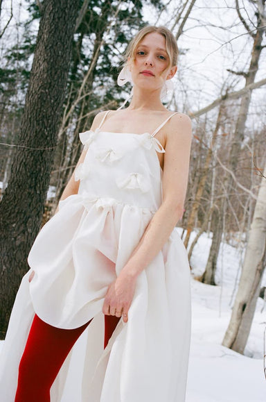 A woman stands outdoors in a snowy forest, wearing the Ariel Dress — Bridal Edit by Noémiah, a silk organza dress with bow details and Swarovski crystals. Bright red tights contrast the white dress against the wintry scene.