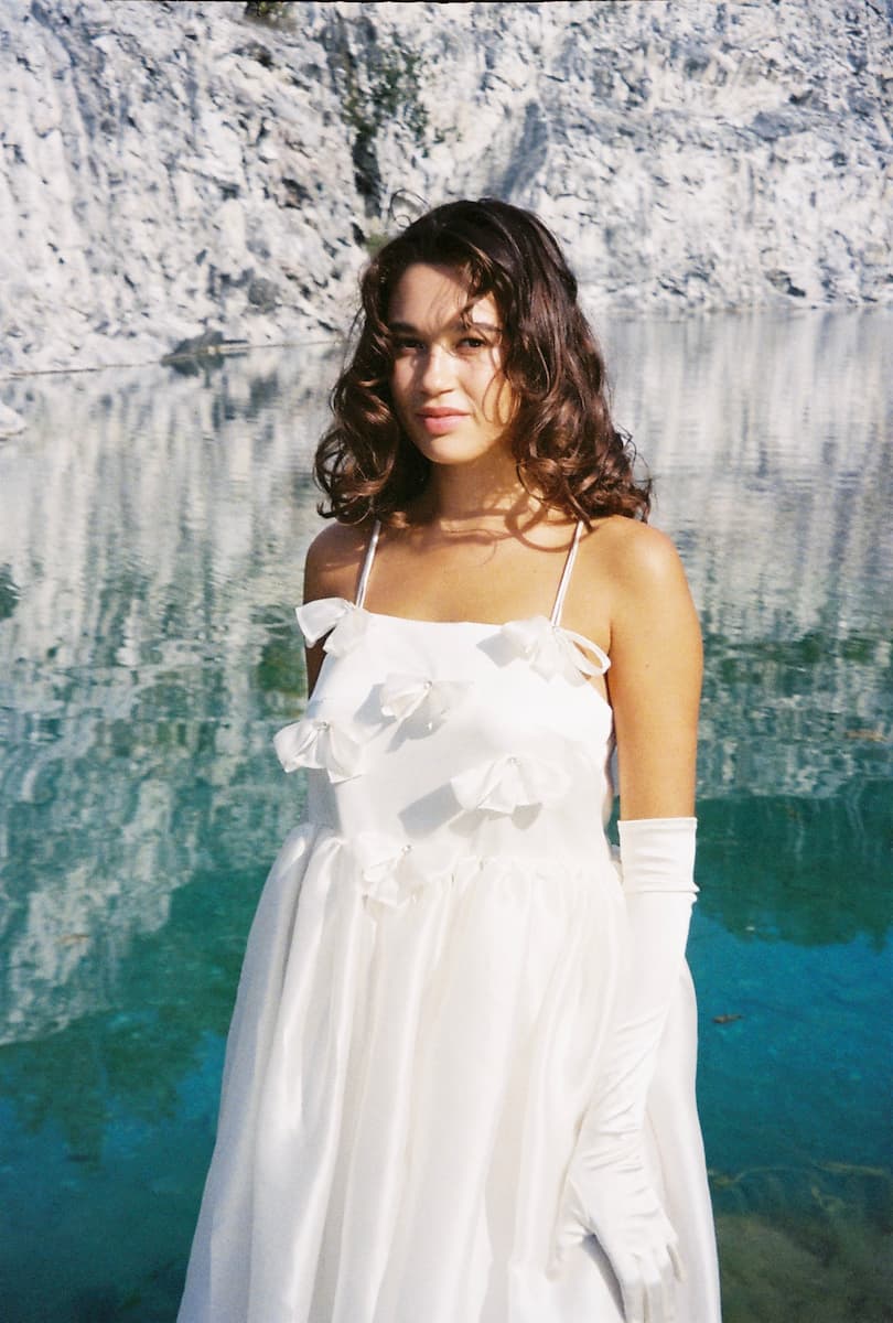 A woman wearing the Noémiah Ariel Dress — Bridal Edit, adorned with Swarovski crystals and long white gloves, stands by clear turquoise water and white cliffs, gazing gently at the camera.