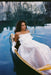 A woman in Noémiah’s Ariel Dress — Bridal Edit, a white silk organza gown with delicate bows, sits thoughtfully in a canoe on calm blue water, her serene gaze mirrored among blurred reflections of trees and sky.