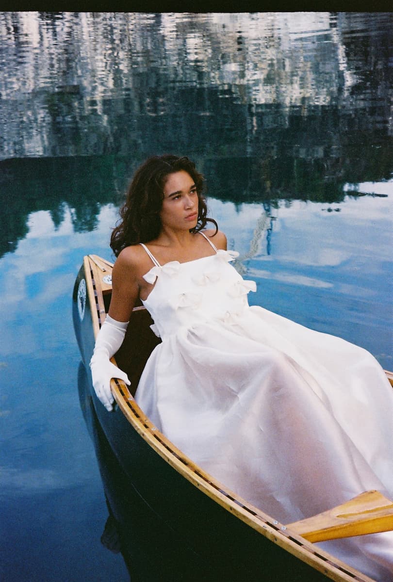 A woman in Noémiah’s Ariel Dress — Bridal Edit, a white silk organza gown with delicate bows, sits thoughtfully in a canoe on calm blue water, her serene gaze mirrored among blurred reflections of trees and sky.