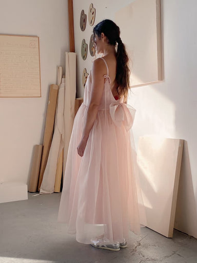 In an art studio, a woman stands in the Noémiah Ariel Dress — Classic, featuring silk organza with a large back bow. Her hair is tied in a ponytail as she gazes at canvases and artwork on the wall, while soft light casts shadows on the floor.