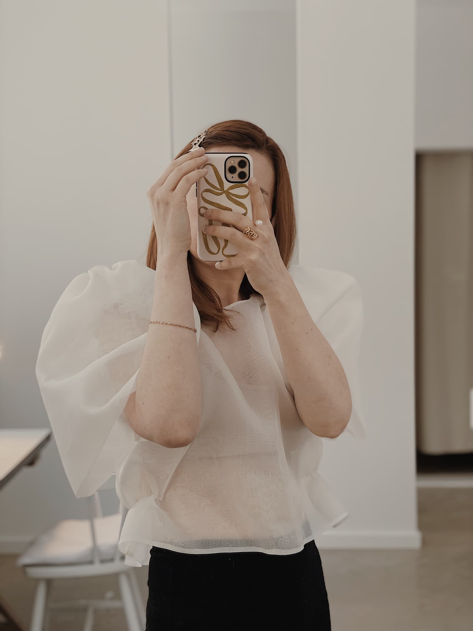 A person with long hair is taking a selfie using a smartphone in front of a mirror. They are dressed in an Arlette Top by Noémiah, which has puffy sleeves, paired with a dark skirt. The room boasts a minimalist design, with a chair and table visible in the background.