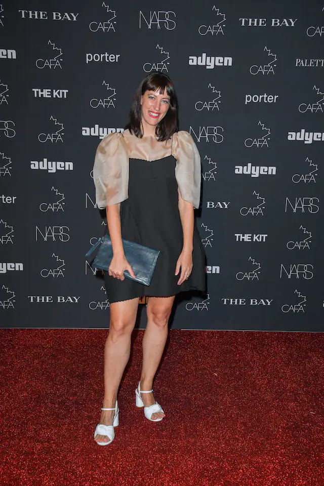 A woman in an Arlette Top featuring sheer puffed sleeves poses on the red carpet. Holding a clutch, she stands before a backdrop adorned with logos like NARS and CAFA. Her silk organza ensemble is complemented by white heels, and she beams with elegance. The top is designed by Noémiah.