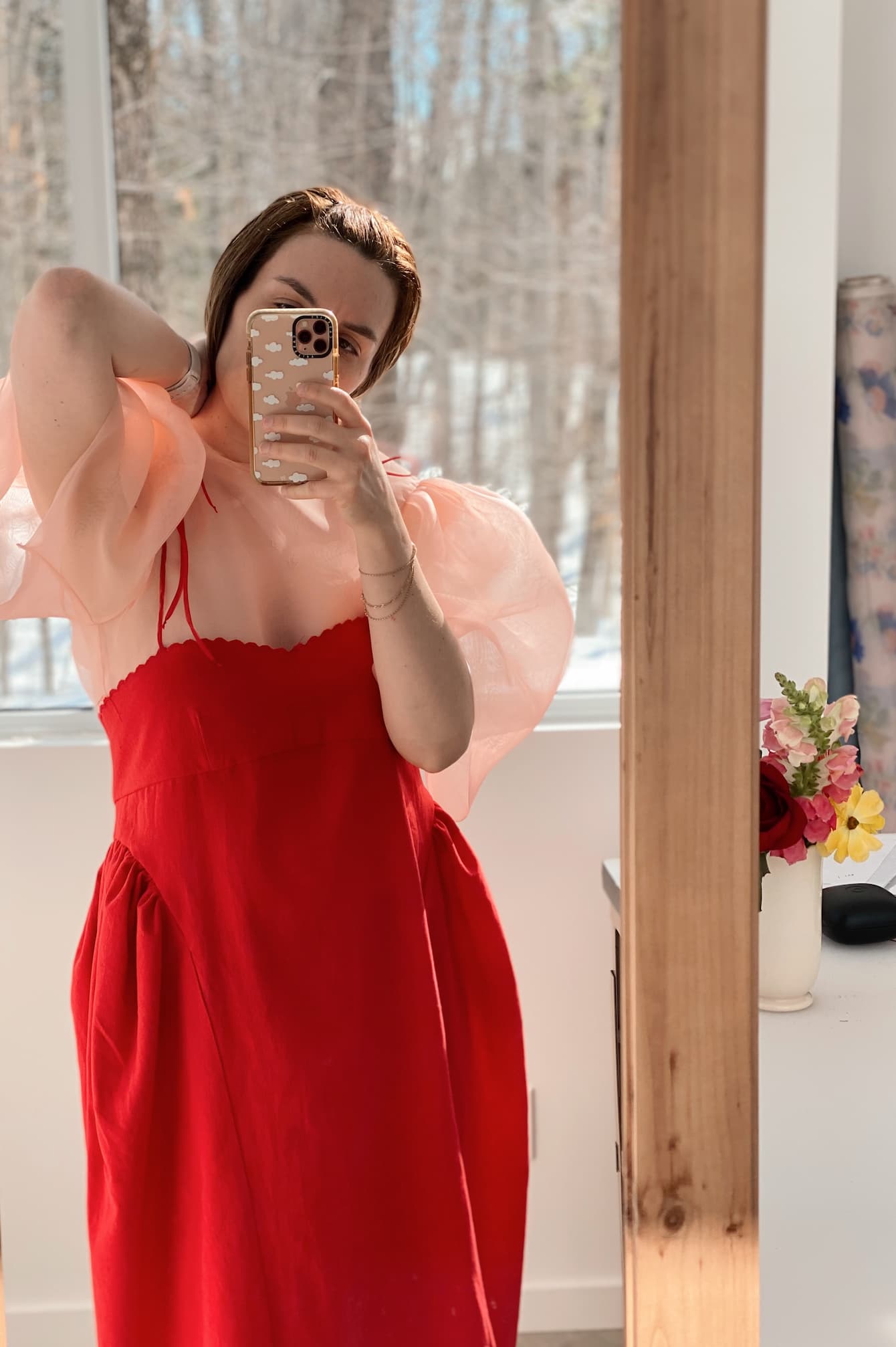 A person wearing an Arlette Top by Noémiah, featuring puffy pink sleeves, takes a mirror selfie. They hold a phone with a decorated case, while in the background there's a vase of flowers on the table and a view of trees outside the window.