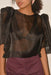 A person wearing a sheer black Arlette Top by Noémiah, featuring puffed sleeves and an intricate pattern, paired with high-waisted purple trousers. The person's hair is styled in curls against a neutral, light background.