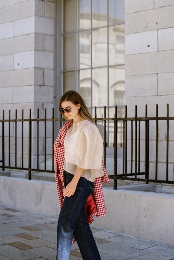 A woman strolls by a stone building in her Arlette Top and sunglasses. Draped over her arm is a red and white checkered coat. The building's large barred windows cast shadows on her as she passes by.