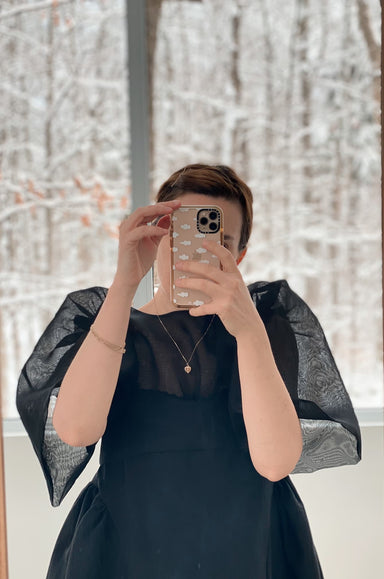 The person is taking a selfie in front of a window, dressed in an impressive Arlette Top by Noémiah with sheer, patterned sleeves resembling silk organza. They're holding a phone with an ornate case, and behind them stretches a picturesque snowy landscape.