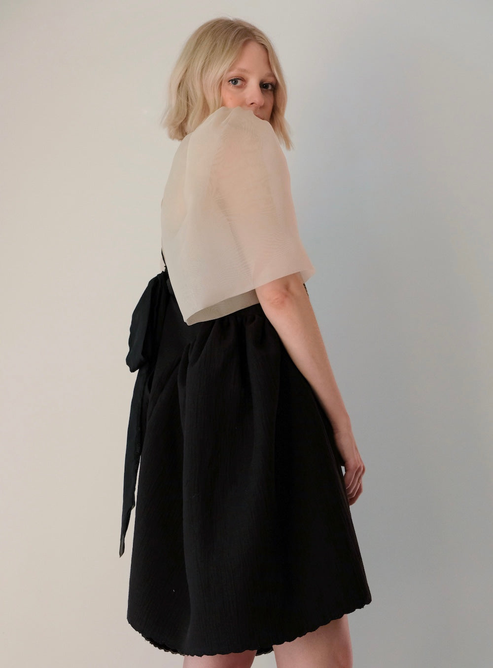 A person with blonde hair is wearing a sheer beige Noémiah Arlette Top along with a black Capucine dress featuring a prominent black bow at the back. Made from silk organza, they stand against a plain light-colored background, glancing over their shoulder.