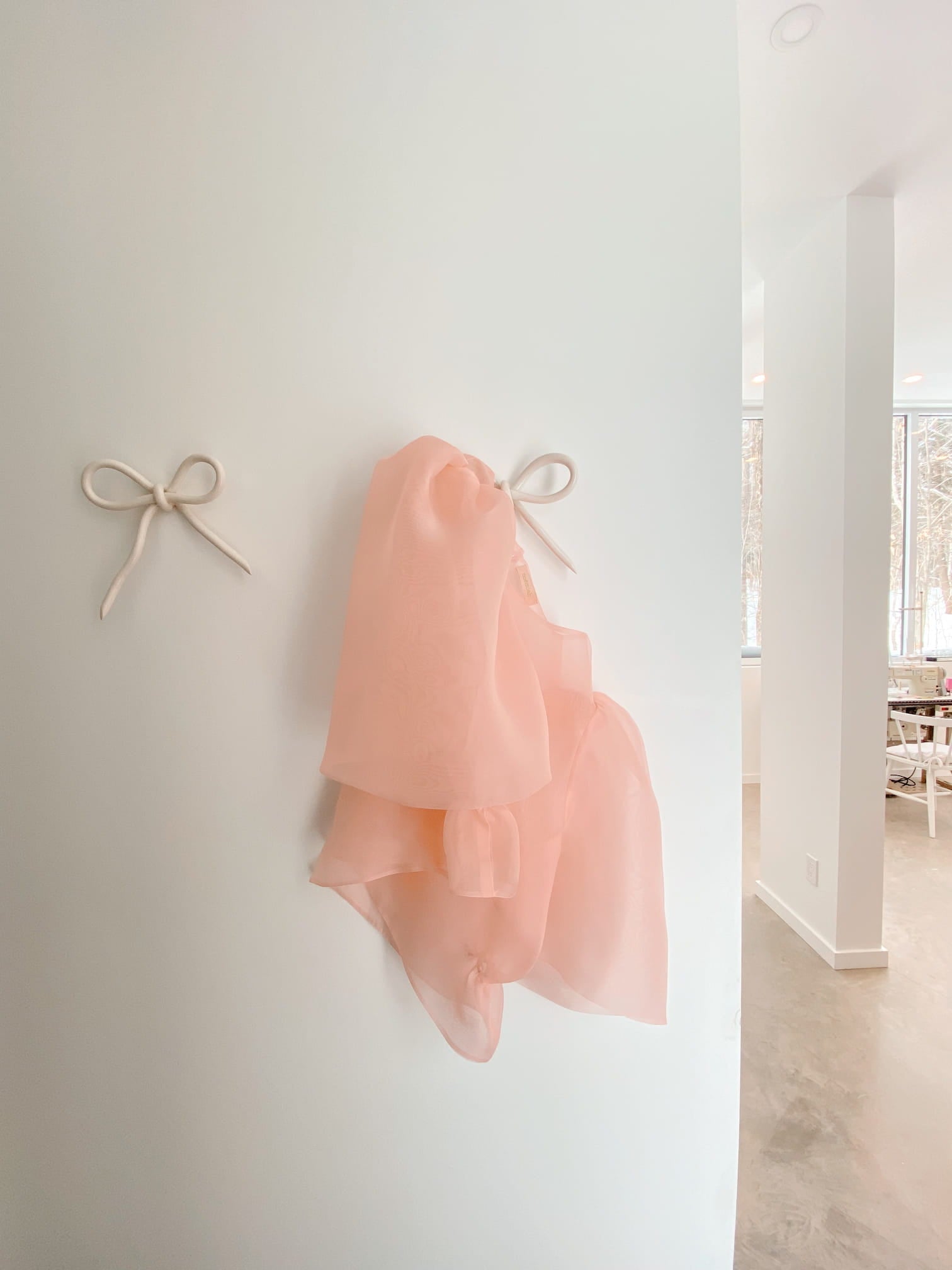 An Arlette Top by Noémiah, made from delicate pink silk organza, is displayed on a white wall from a decoratively designed, bow-shaped hook, with an identical empty hook beside it. The background reveals a bright room featuring large windows and part of a table.