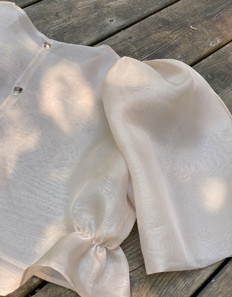 The Arlette Top by Noémiah, crafted from sheer silk organza with long sleeves and gathered cuffs, rests elegantly on a wooden surface, its button details shining as the soft sunlight casts gentle shadows.