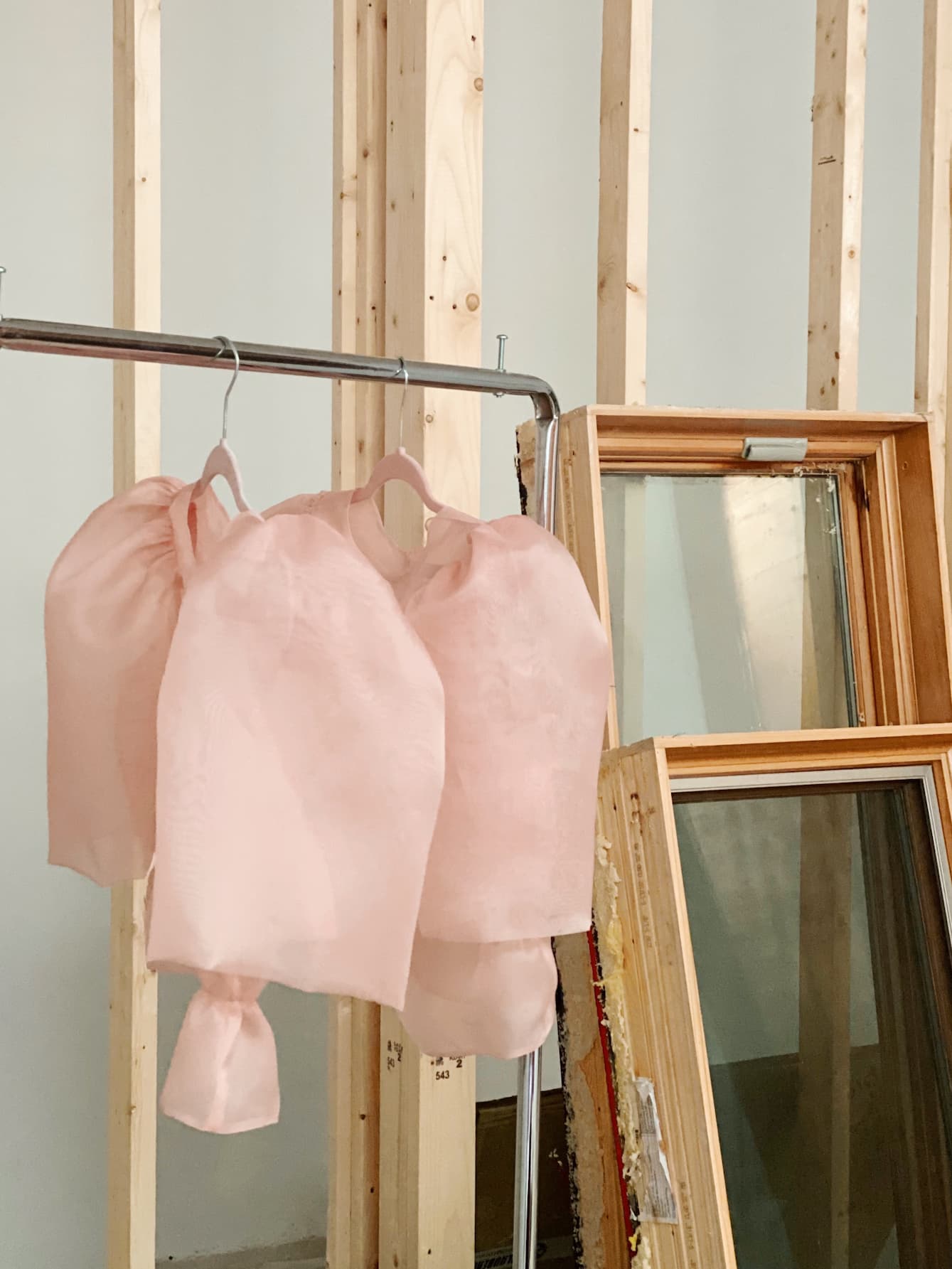 A wooden frame structure featuring two pastel pink fabric pieces, reminiscent of an Arlette Top by Noémiah, hangs gracefully on a metal rack. Next to it, several wooden-framed mirrors lean casually against the wall, reflecting elegance and style.