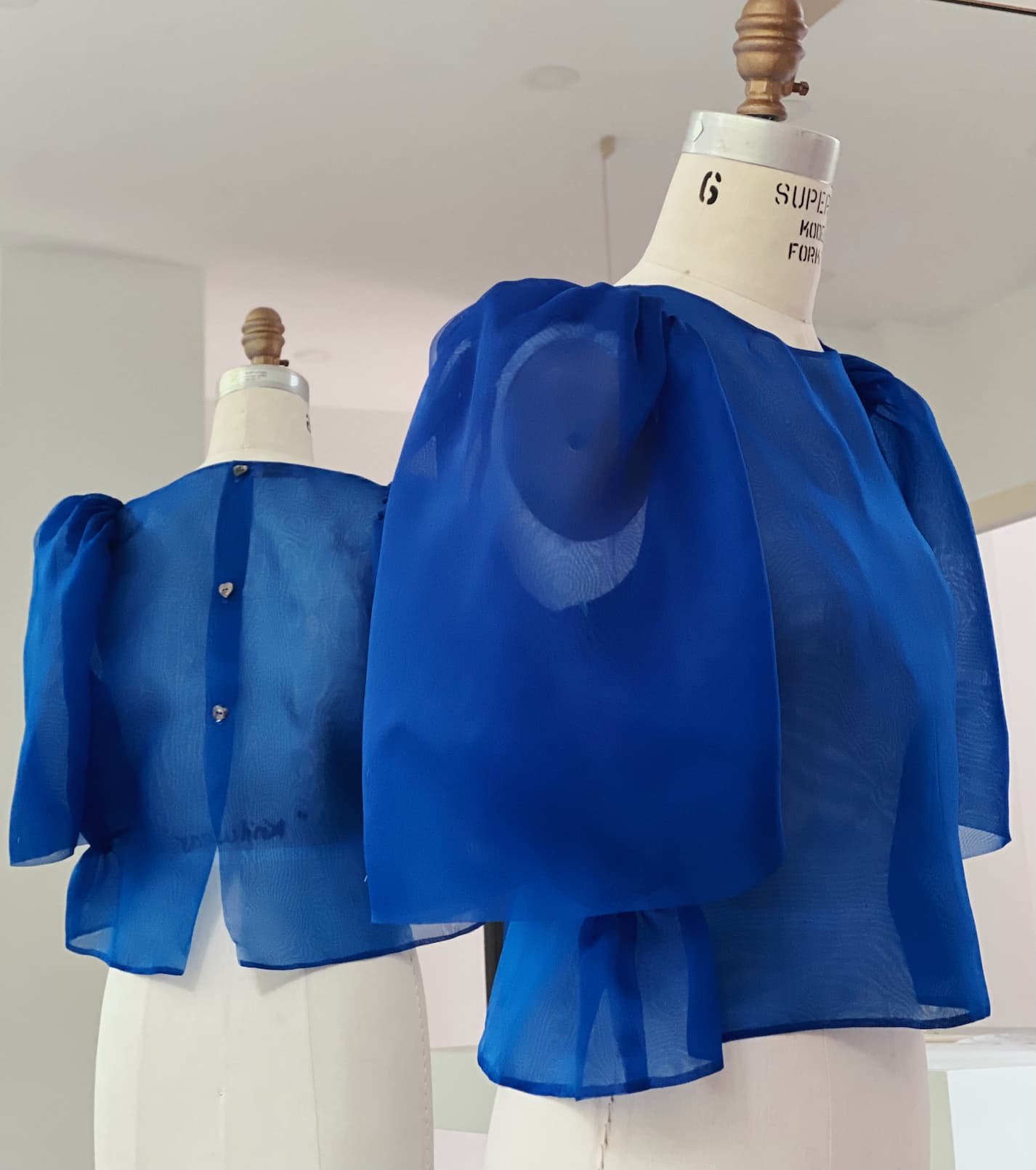Two dress forms display the Noémiah Arlette Top—a sheer royal blue silk organza piece with voluminous puffed sleeves. One shows the front; the other reveals button detailing at the back, set in a minimal white indoor space. Plus size available.