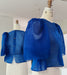 Two dress forms display the Noémiah Arlette Top—a sheer royal blue silk organza piece with voluminous puffed sleeves. One shows the front; the other reveals button detailing at the back, set in a minimal white indoor space. Plus size available.