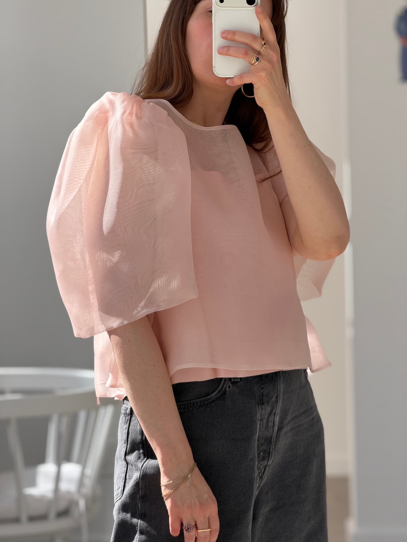 Wearing the Arlette Top by Noémiah—a sheer, pale pink silk organza blouse with puffed sleeves—and dark high-waisted jeans, a person takes a mirror selfie with their phone in a sunlit room.