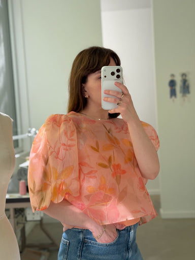 A person wears an Arlette Top — Floral Edit in peach silk organza with puffed sleeves and an orange floral pattern, tucked into blue jeans.