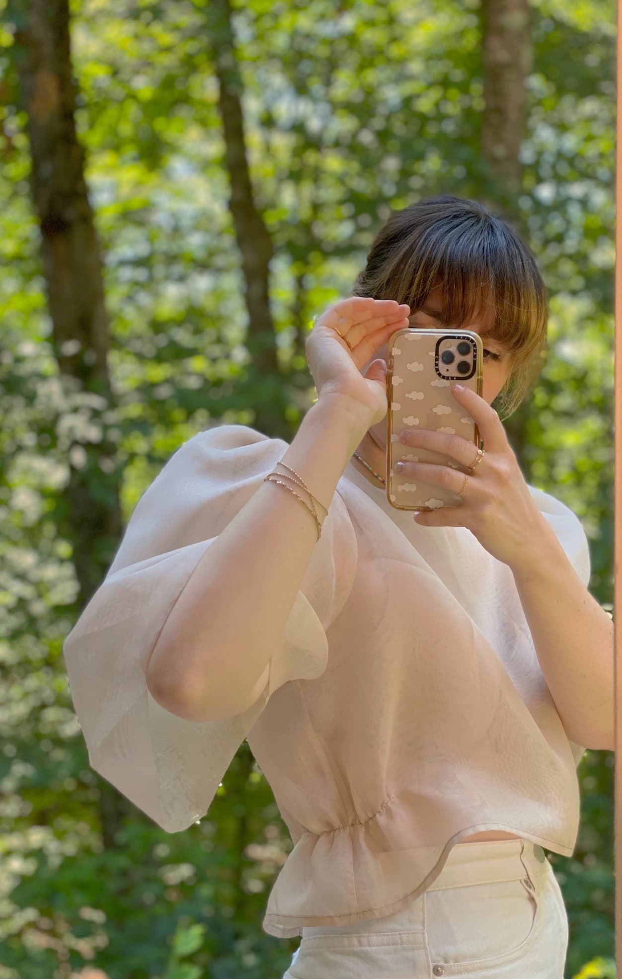 A person with bangs captures a mirror selfie in the forest, wearing a light, puff-sleeved Arlette Top by Noémiah and white pants. The backdrop features lush green foliage, setting a serene outdoor scene.