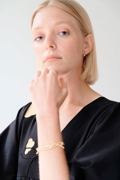 A person with short blond hair is looking at the camera with a neutral expression. They are wearing a black top with gold accents, complemented by a gold-plated drop bracelet. The refined look is completed with Béatrice Earrings from Noémiah and the simple white wall in the background.