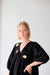 A person with short blond hair wears a black dress adorned with gold cloud embellishments and pairs it with Béatrice Earrings by Noémiah. They stand against a plain white background, looking at the camera with a neutral expression.