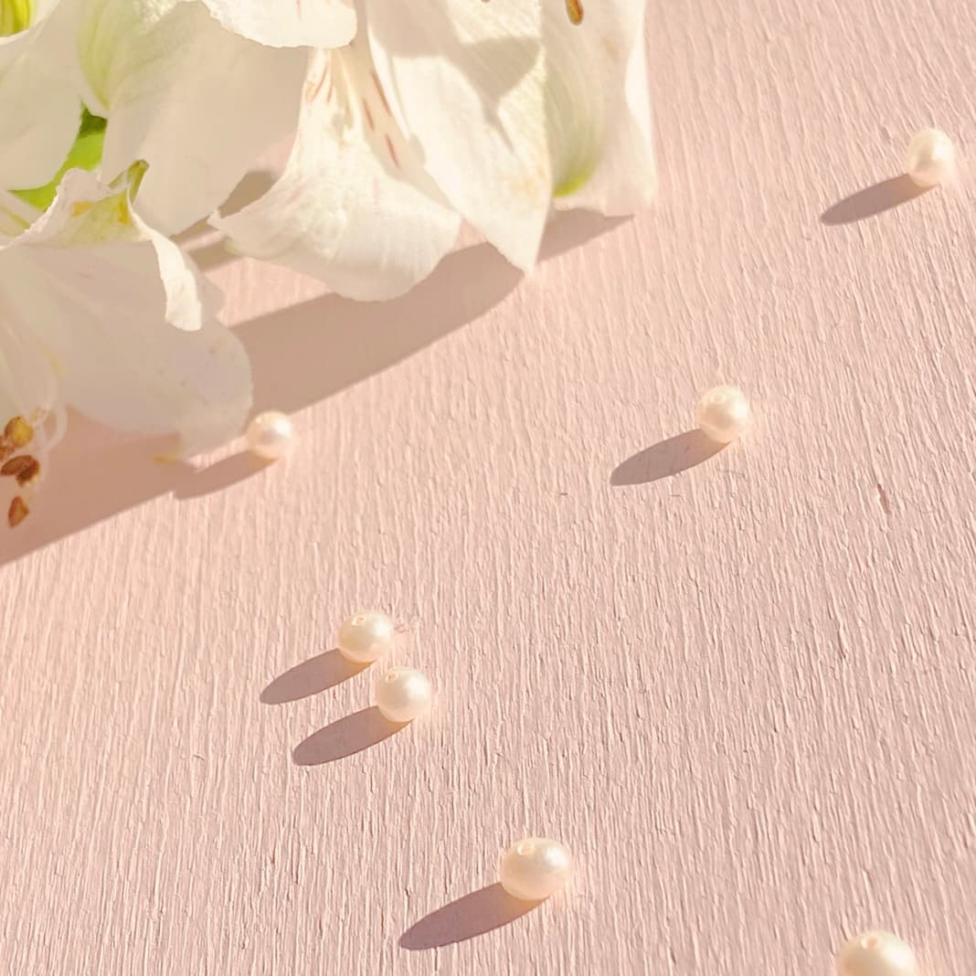 The Béatrice Earrings by Noémiah, adorned with white flowers and freshwater natural pearls, are artfully arranged on a soft pink surface, resulting in a serene and elegant composition accentuated by gentle shadows.