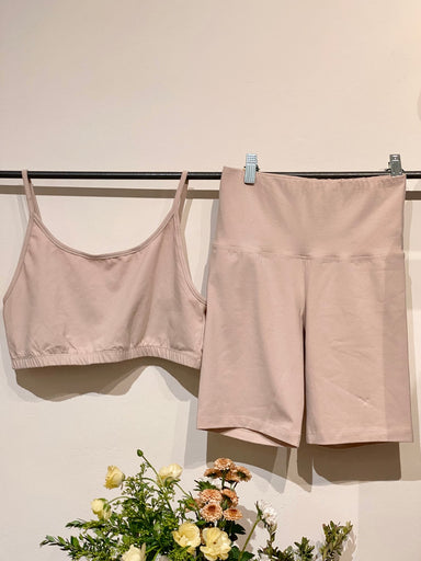 A beige sports bra paired with Noémiah's Bike Shorts is displayed on a rack against a plain wall. Underneath, a vibrant bouquet of assorted flowers enhances the scene with a splash of color. The set, made from organic cotton, embodies stylish and sustainable fashion choices.