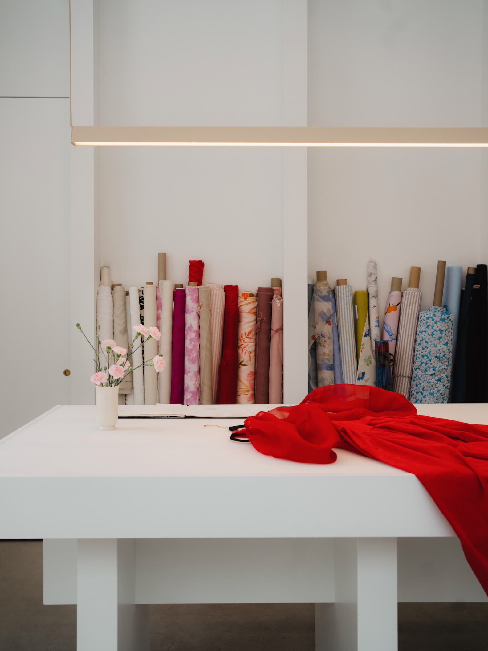 In the serene sewing studio of Noémiah, a white table is elegantly draped with sleek silk organza, while a mug holding delicate pink flowers sits on top. Behind, rolls of vibrant fabric stand ready to be transformed into the Billie Dress from Noémiah's collection, neatly arranged on shelves against an immaculate white wall.