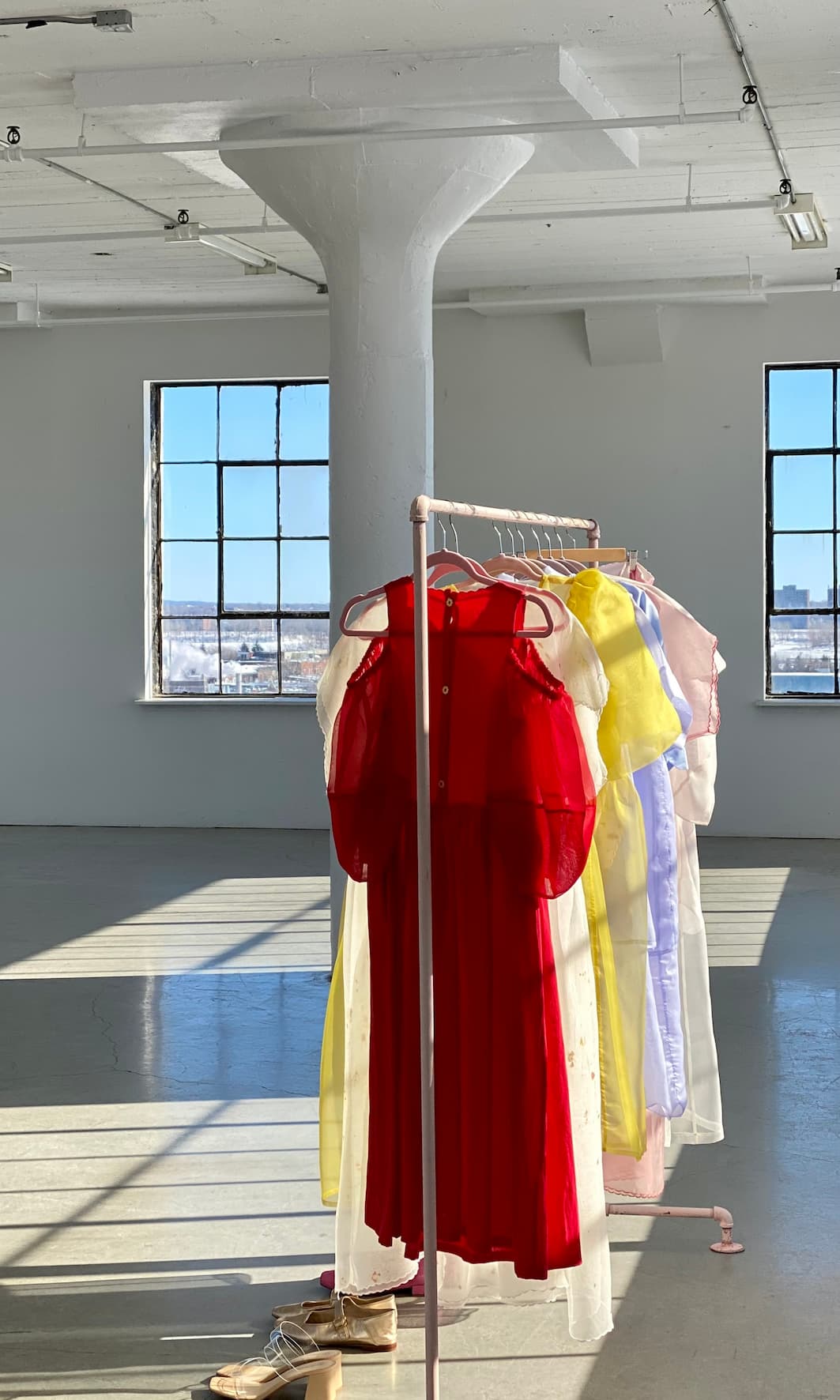 A sunlit room with large windows and a concrete column features a clothing rack displaying several elegant Billie Dresses by Noémiah, made in vibrant red, yellow, and pastel silk organza. A pair of shoes rests on the floor beside the rack.