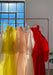 A clothing rack displays five sheer dresses by Noémiah in white, yellow, peach, pink, and red. Sunlight filters through the large window behind them, casting soft shadows in a bright, minimalist setting.