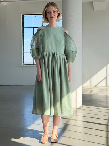 A woman with short blonde hair smiles slightly indoors by a window, wearing the Noémiah Billie Dress—a light green crumple cotton dress featuring sheer silk organza puff sleeves and cold-shoulder cutouts.