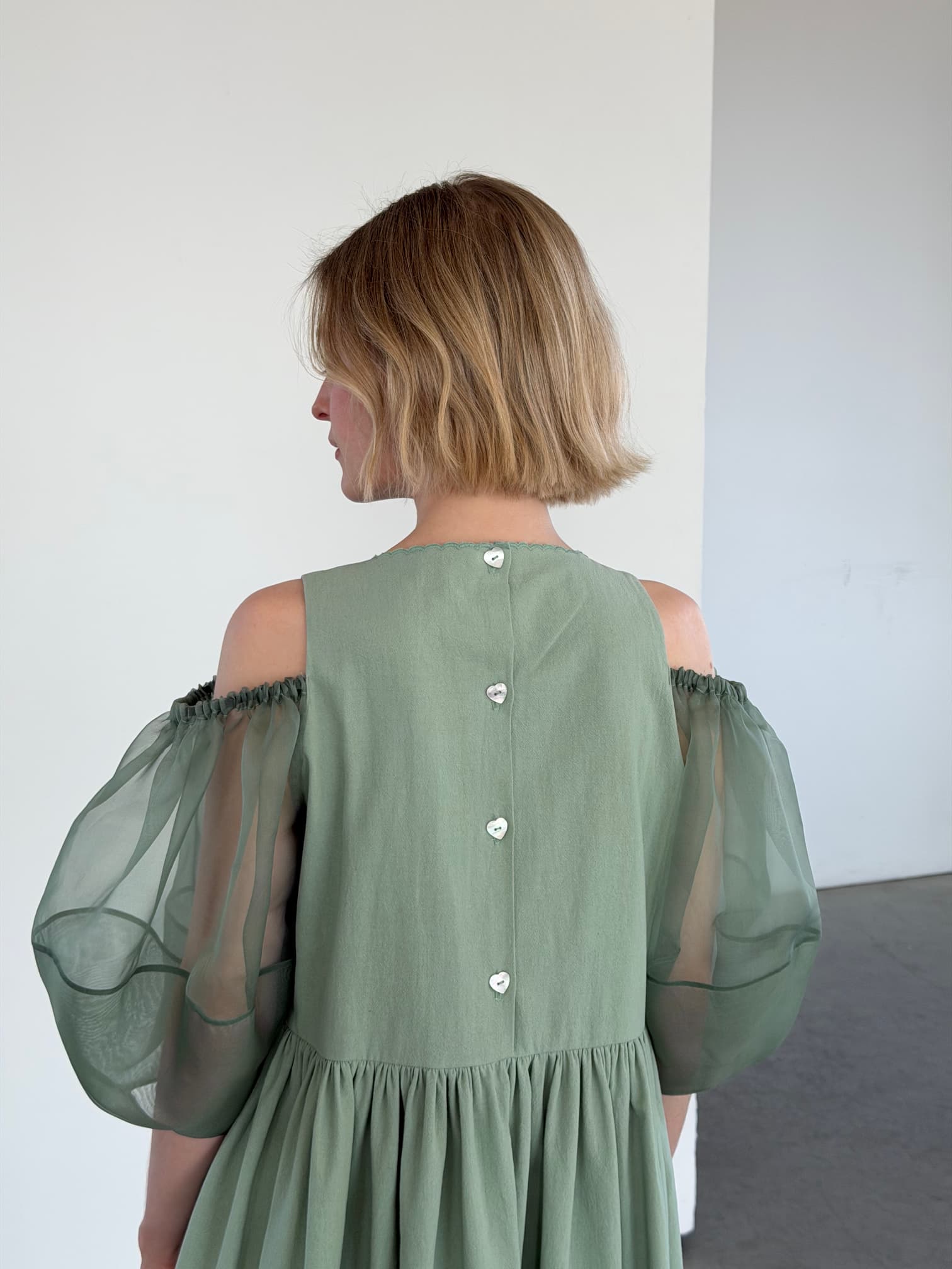 A woman with short blonde hair is seen from behind wearing the Noémiah Billie Dress—sage green crumple cotton with silk organza sleeves and decorative back buttons—in a minimalist, light-filled room with white walls.