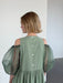 A woman with short blonde hair is seen from behind wearing the Noémiah Billie Dress—sage green crumple cotton with silk organza sleeves and decorative back buttons—in a minimalist, light-filled room with white walls.