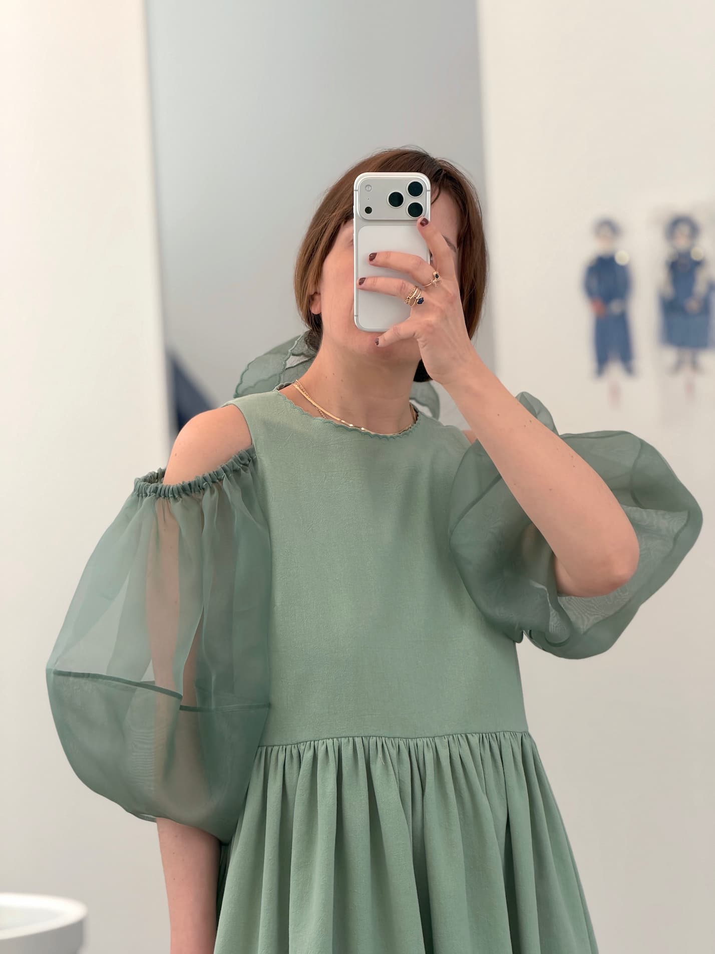 A person wears the light green Billie Dress by Noémiah, featuring crumple cotton and sheer silk organza puffy sleeves, taking a mirror selfie that covers their face. The minimal background has two small wall illustrations.
