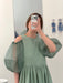 A person wears the light green Billie Dress by Noémiah, featuring crumple cotton and sheer silk organza puffy sleeves, taking a mirror selfie that covers their face. The minimal background has two small wall illustrations.