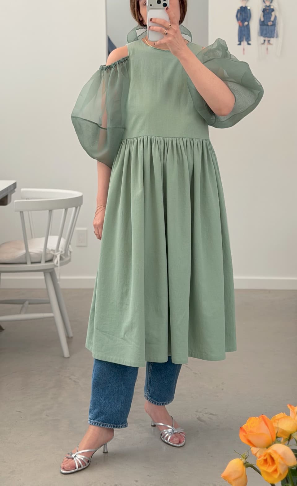 A person takes a mirror selfie wearing the sage green Billie Dress by Noémiah with off-shoulder, puff-sleeve details, styled over blue jeans and silver heels. Flowers and a white chair are visible in the room.