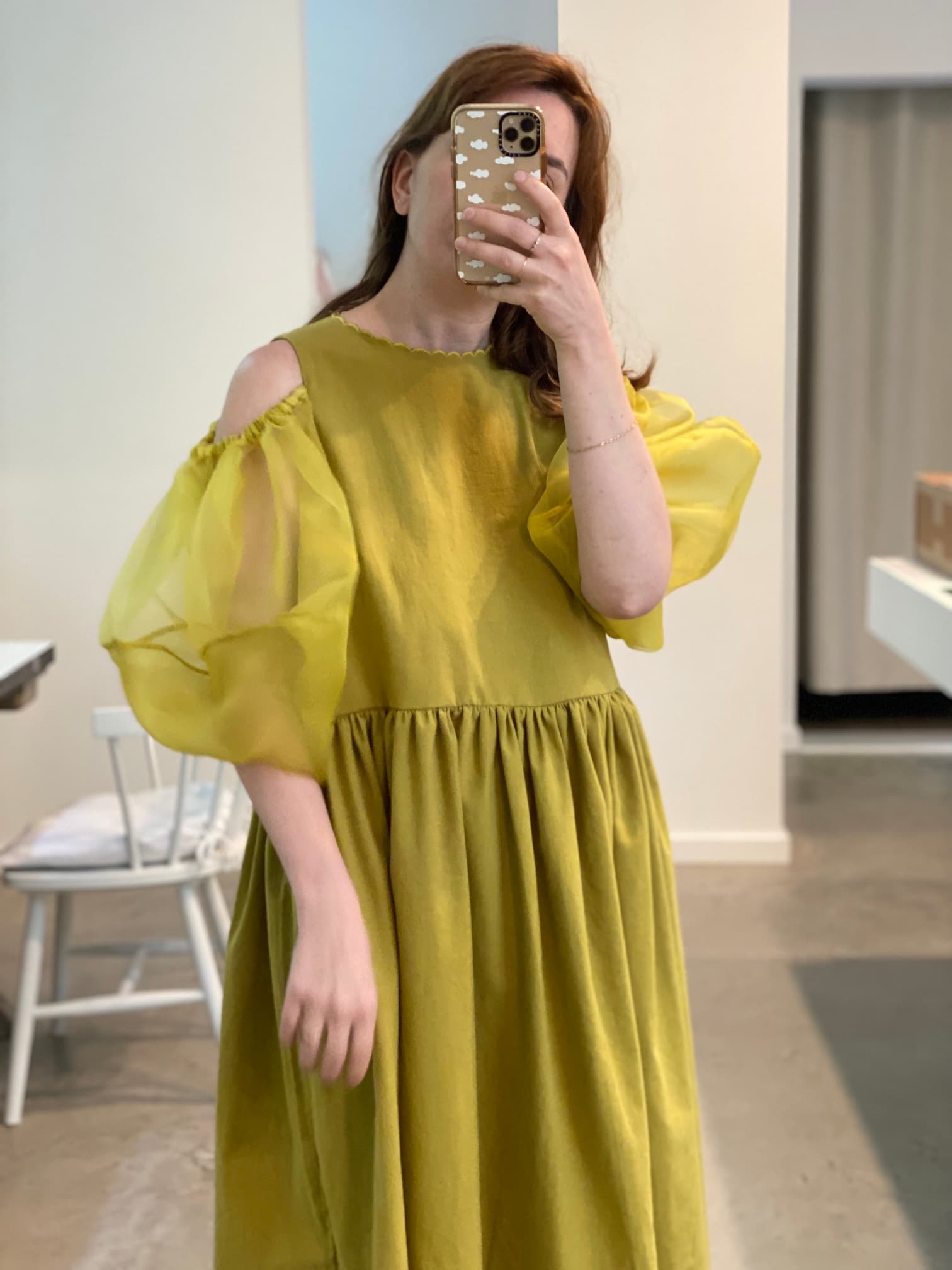 A person takes a mirror selfie wearing the Billie Dress by Noémiah—an elegant green, long-sleeved dress featuring sheer, puffy shoulders crafted from silk organza and a gathered waist. The scene unfolds in a minimalistic room with white walls and a chair.