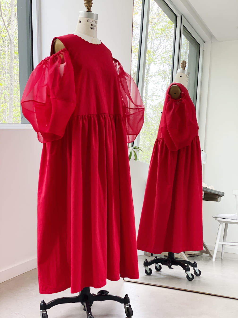 The Billie Dress from Noémiah, with its elegant red color and sheer puff sleeves, is showcased on a mannequin in a well-lit room with large windows. The silk organza fabric provides a touch of sophistication as the dress is reflected in a mirror, highlighting its loose, flowing design.