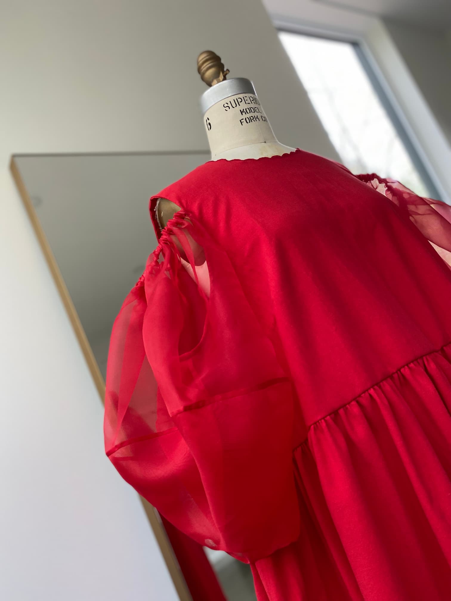 The Billie Dress by Noémiah, an elegant red creation crafted from silk organza with sheer puff sleeves, is displayed on a mannequin. The backdrop features a large mirror and a window that illuminates the scene with natural light.