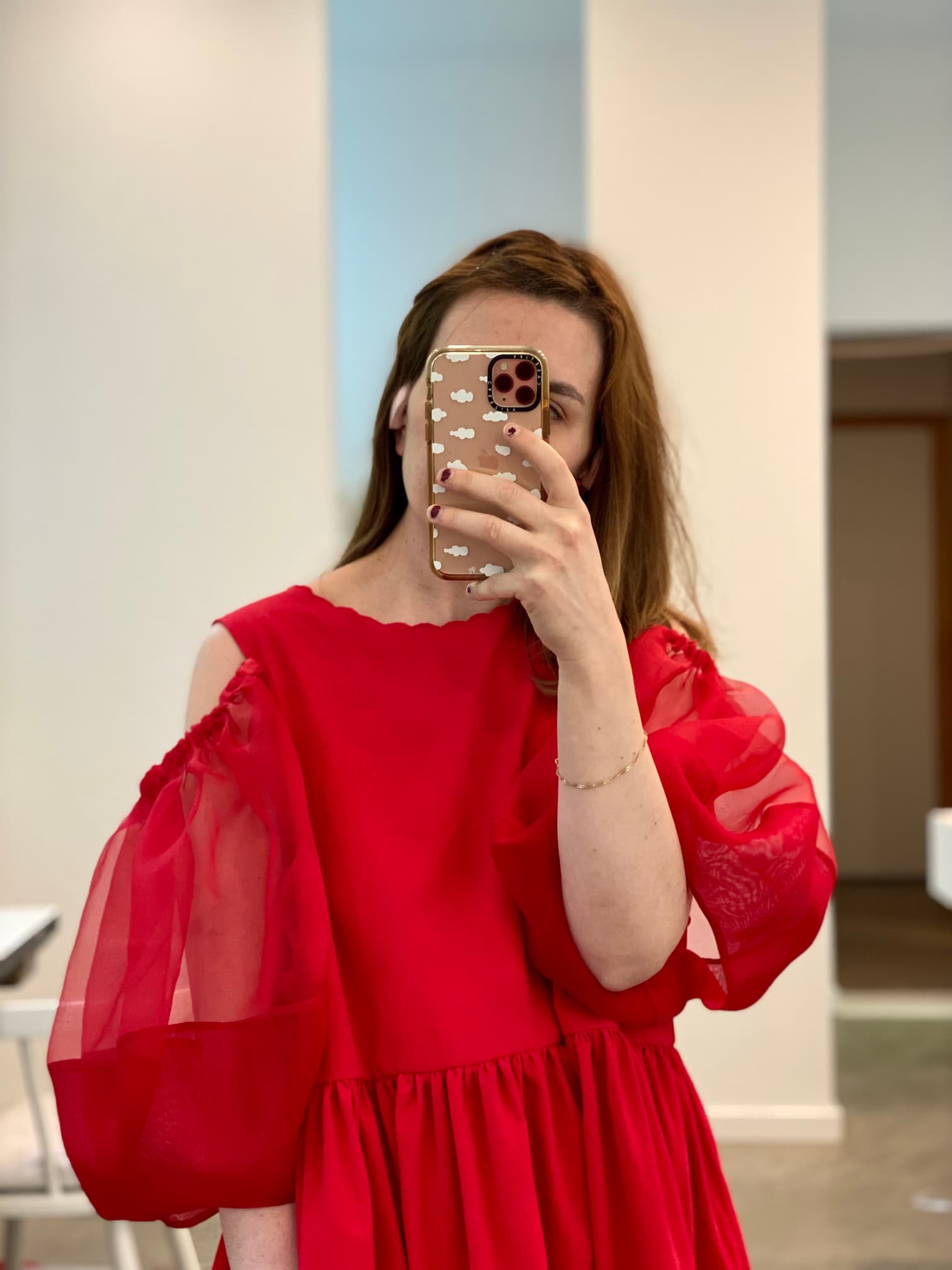 A person with long hair wearing the Noémiah Billie Dress, featuring elegant sheer sleeves, takes a mirror selfie. They hold a phone encased in a cloud-patterned design, standing in a well-lit room adorned with white and wooden elements that exude timeless charm.