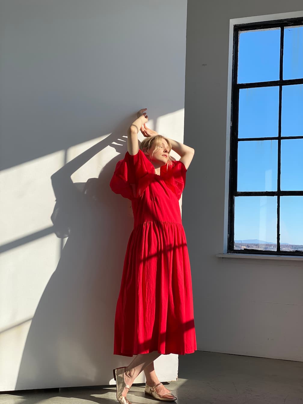 A person in the elegant, flowing Billie Dress by Noémiah poses gracefully against a sunlit white wall. Their shadow is cast on the surface, while a large window reveals a clear blue sky outside. The silk organza fabric of the dress catches the light beautifully, adding to the scene's allure.