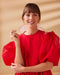 A smiling woman stands against a beige background, wearing the elegant Billie Dress by Noémiah, featuring puffed sleeves. Crafted from silk organza, it complements her bangs and statement earrings. With her left hand close to her face, she exudes a calm and confident demeanor.