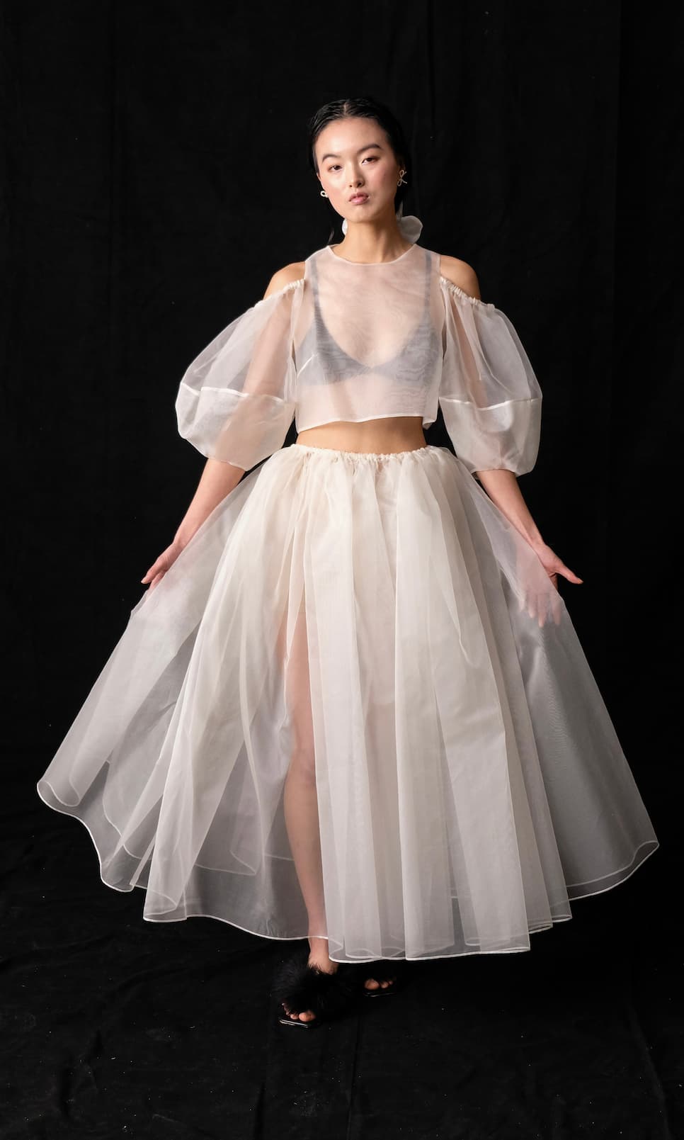 A person stands against a black background wearing the Billie Top by Noémiah, a sheer, white bridal ensemble with puffy, off-shoulder lantern sleeves. They pair it with a long, flowing skirt and style their hair back, complemented by black footwear.