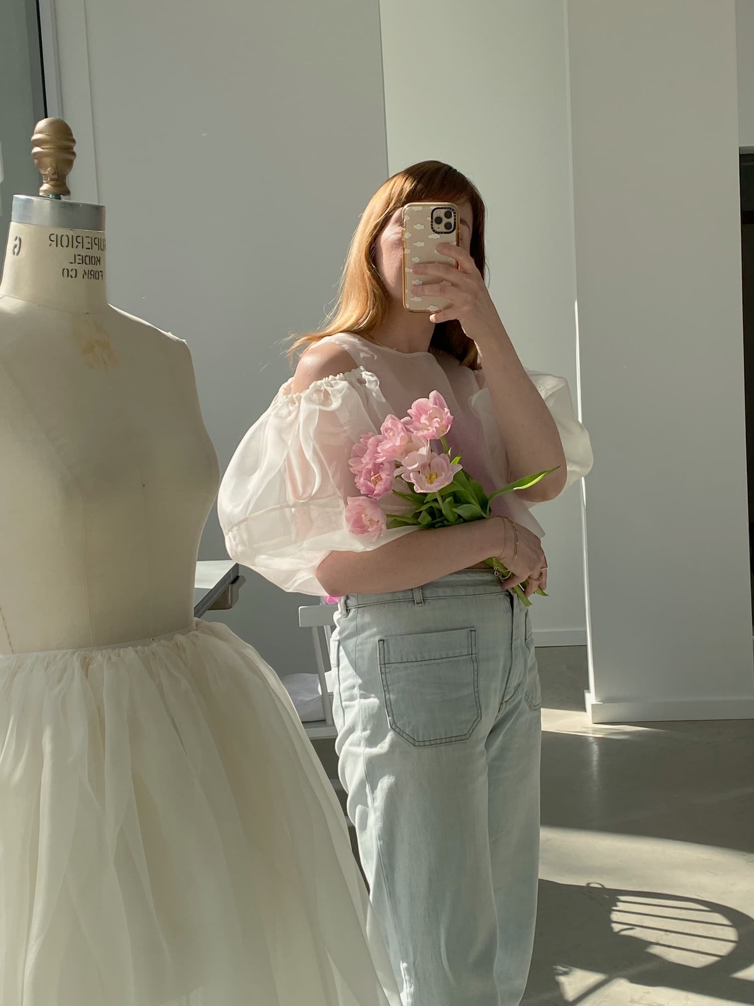 A person with long hair takes a mirror selfie while holding pink flowers, donning the Noémiah Billie Top—a white, off-the-shoulder blouse with delicate lantern sleeves—and light jeans. Beside them stands a dress form adorned with a silk organza tutu. Sunlight streams through the large windows, brightening the room.