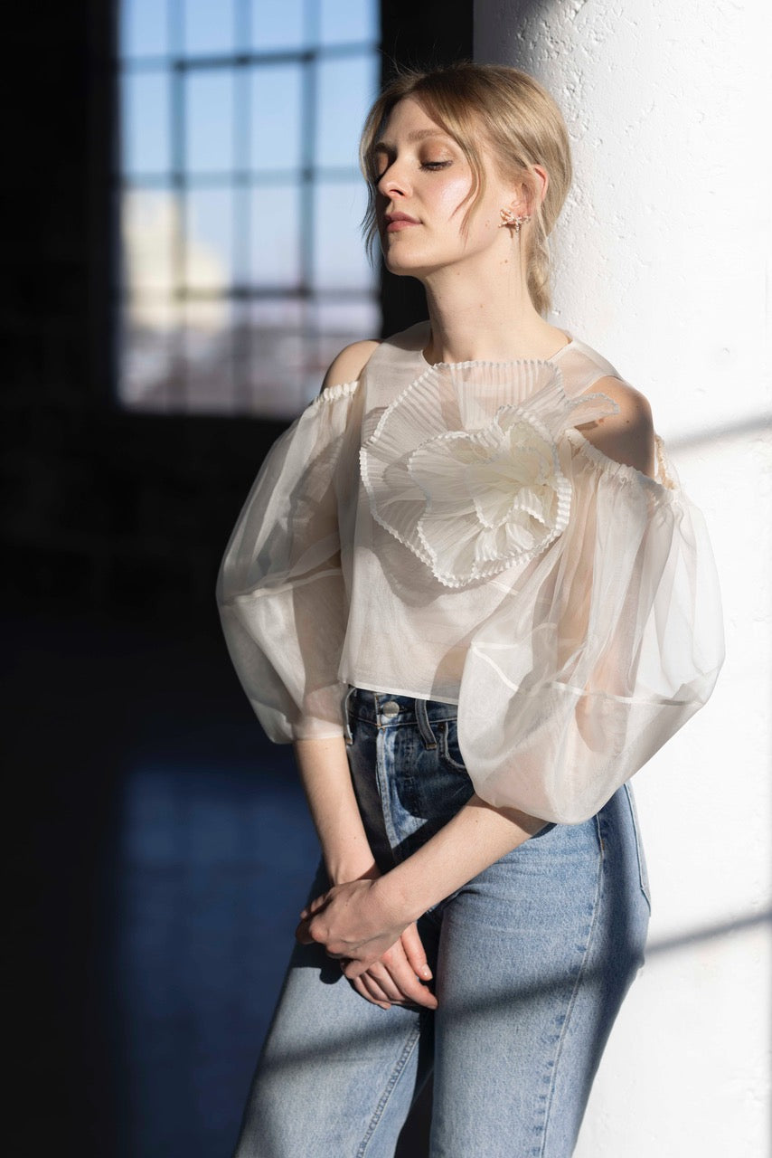 A woman with blonde hair leans against a white pillar in a sunlit room, wearing the Billie Top by Noémiah—a sheer silk organza blouse with lantern sleeves and floral embellishments—paired with blue jeans. Her eyes are closed as the soft, warm light highlights her profile.