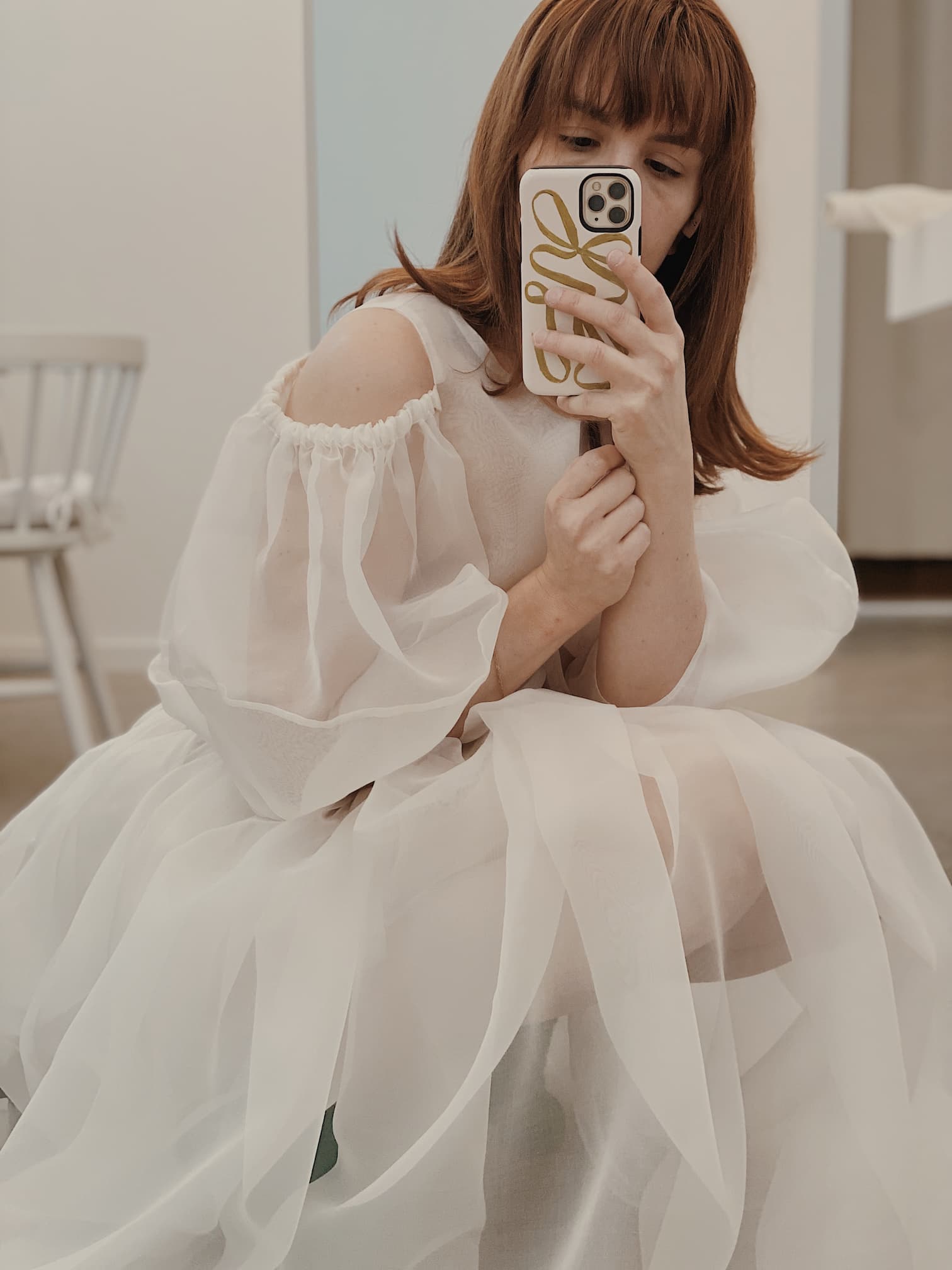 A person with auburn hair is seated, capturing a mirror selfie in the Billie Top from Noémiah. The white sheer design features elegant off-shoulder and lantern sleeves. Their phone case showcases a stylish abstract pattern, set against a backdrop of a white chair and neutral-colored walls.