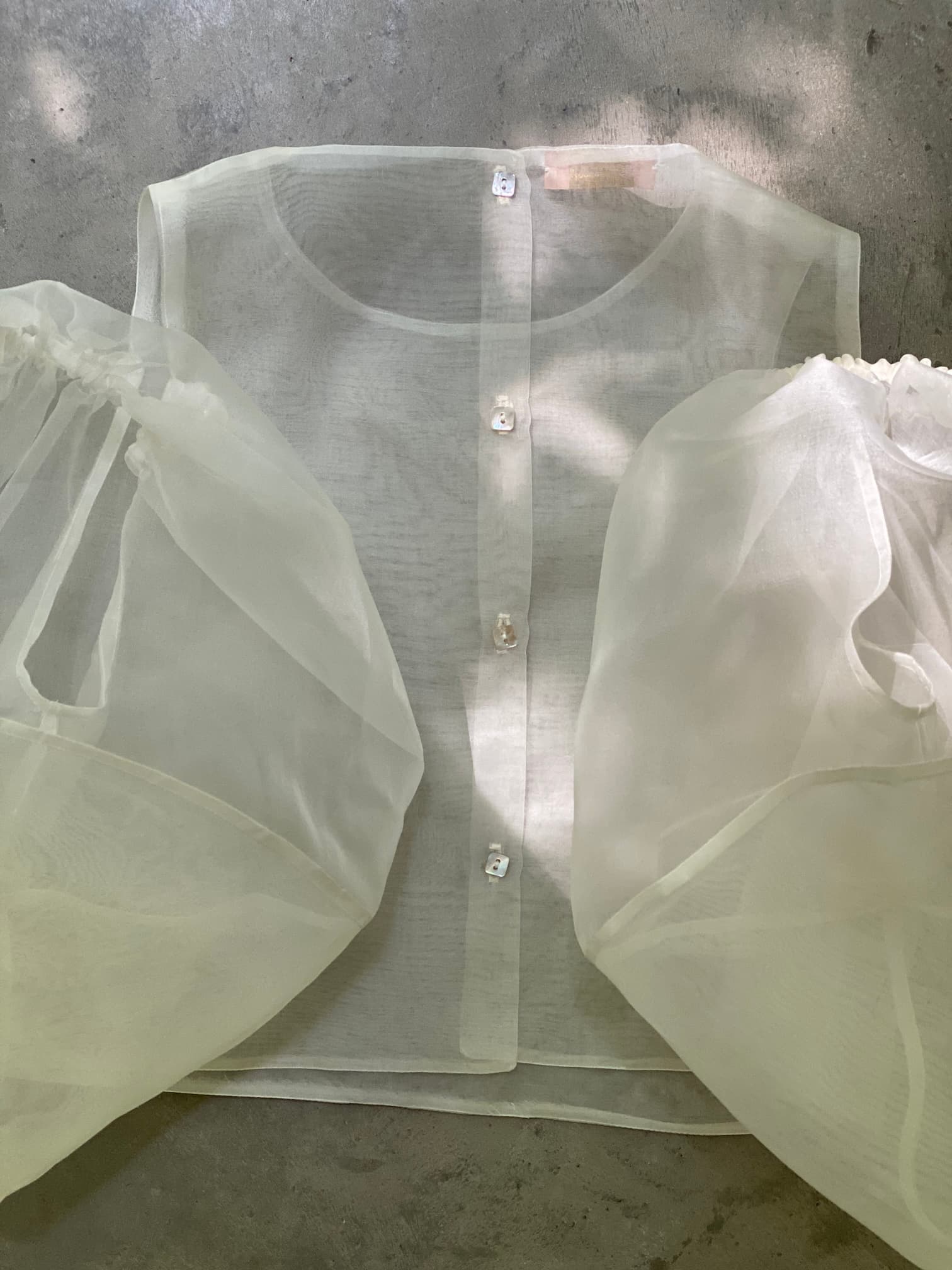 The Billie Top by Noémiah, a sheer button-up blouse with delicate lantern sleeves, is laid on a concrete surface. Its silk organza fabric is translucent, showcasing a soft, airy texture. Shadows from surrounding foliage gently fall across the Billie Top.