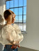 A woman with light hair stands by a wall in a sunlit room, wearing the Billie Top by Noémiah, featuring sheer white fabric and lantern sleeves, paired with blue jeans. She gazes downward as soft sunlight casts a warm glow, with a window in the background.