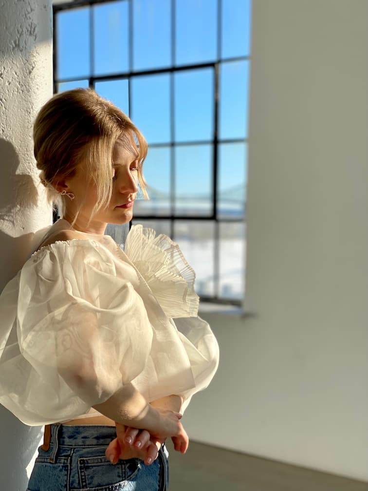 A woman with light hair stands by a wall in a sunlit room, wearing the Billie Top by Noémiah, featuring sheer white fabric and lantern sleeves, paired with blue jeans. She gazes downward as soft sunlight casts a warm glow, with a window in the background.