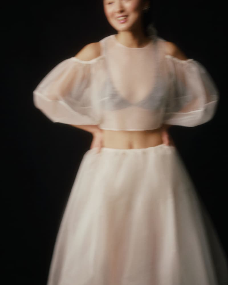 A woman dressed in the Billie Top by Noémiah, a sheer, flowing silk organza piece with lantern sleeves, poses against a dark background. The image is intentionally blurred, creating a dreamy, ethereal effect.