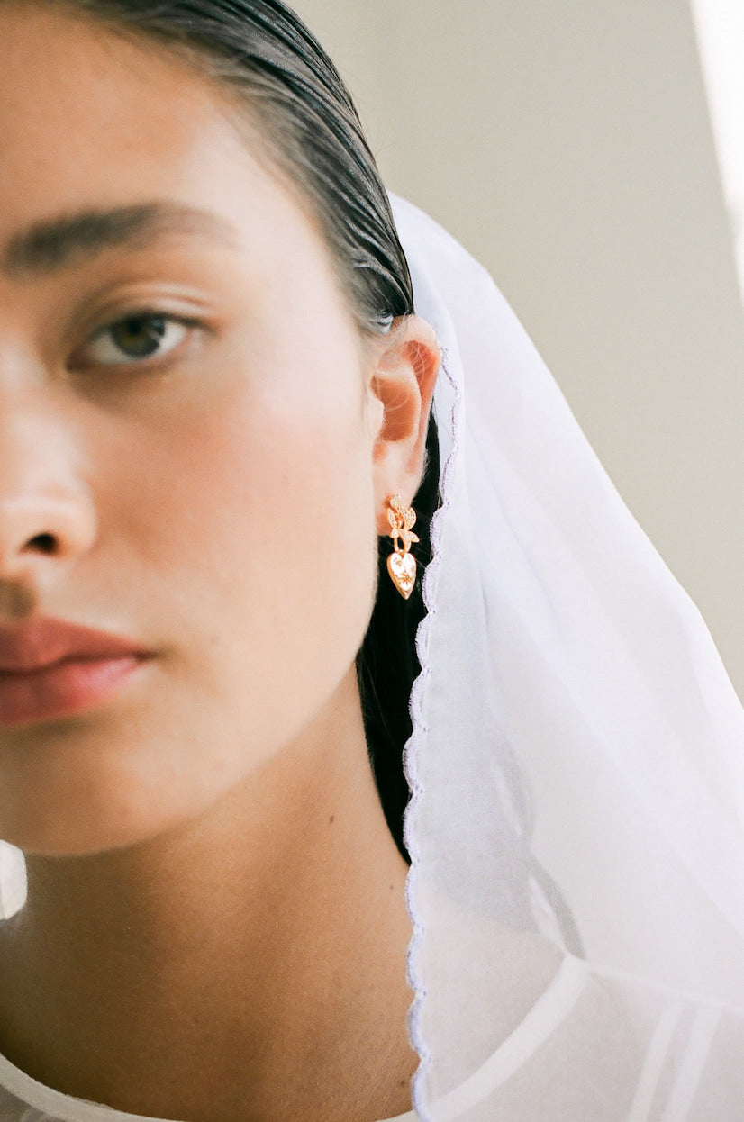 A close-up of a person wearing a white veil and Noémiah's Bouquet Earrings, designed with gold-plated orchid details. The focus is on the left side of their face, highlighting smooth skin and glossy brown hair. Soft natural light creates a serene and elegant ambiance.