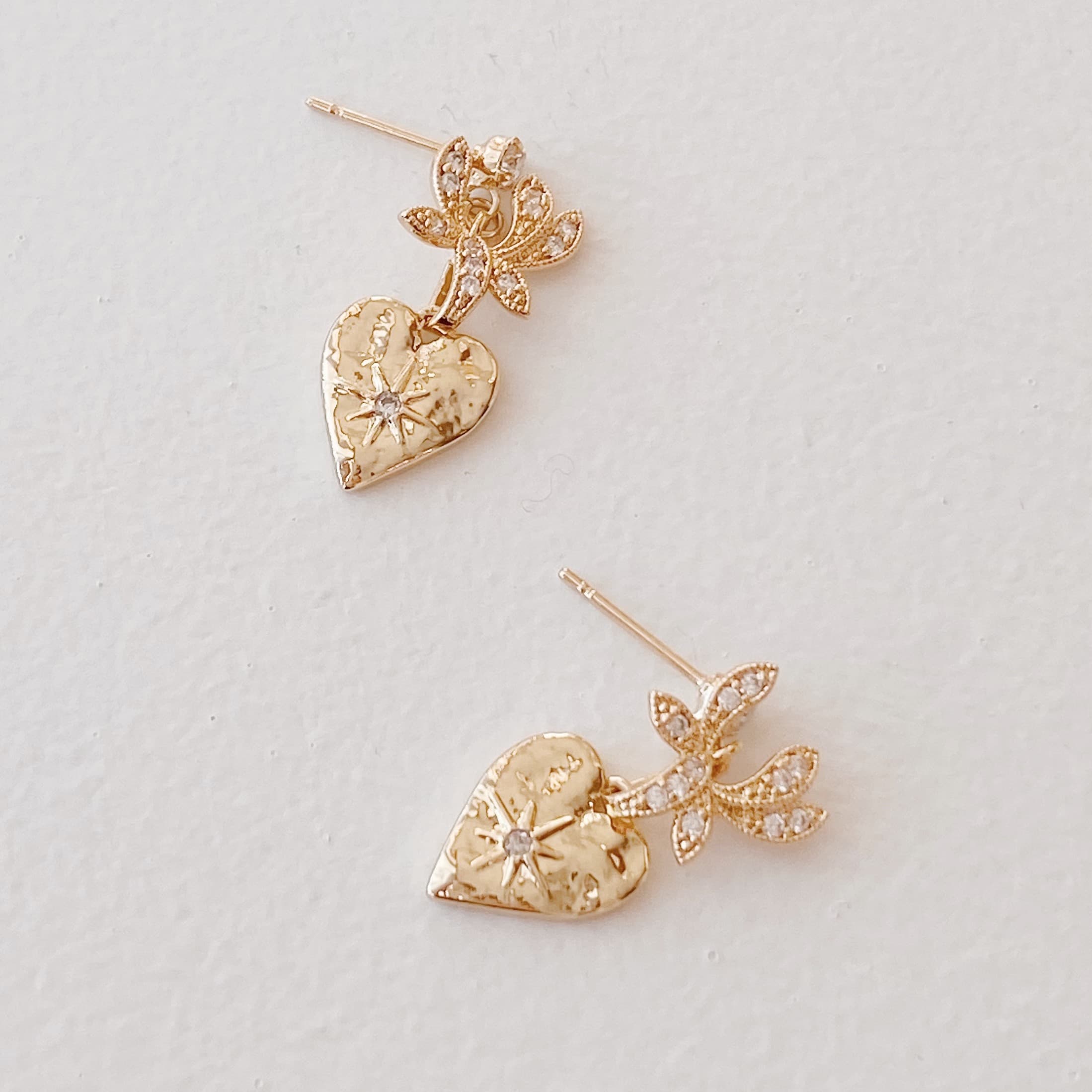 Noémiah's Bouquet Earrings are elegantly showcased on a plain white surface, each featuring a textured heart adorned with cubic zirconia and a decorative floral motif that resembles gold-plated orchids, embellished with tiny sparkling stones.