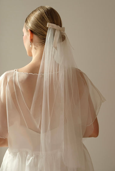 A bride is seen from behind in a sheer wedding dress, with her hair in a sleek bun adorned by Noémiahs Bow Veil, an elegantly placed silk bow securing her long veil. The backdrop is simple and neutral.